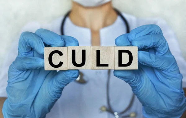 word CULD on wooden cube blocks in the hands in protective gloves of a doctor woman. Blurring doctor on background