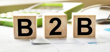 Tahta küplerin dokusunda B2B yazıları var. Hesap makinesi ve iş belgelerinin arka planında tahta küpler var. Finansal, ticari ya da ekonomik tema üzerine bir yazı.