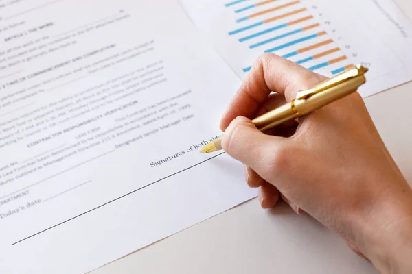 Young business woman signs a business contract on the background of economic charts while sitting at the table. Business and finance concept. Selective focus.