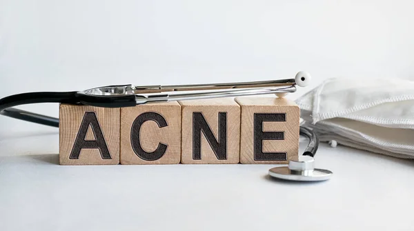 Beyaz arka planda izole edilmiş ahşap küplerdeki ACNE yazıtları, tıp konsepti. Masanın yanında steteskop ve koruyucu maskeler var..