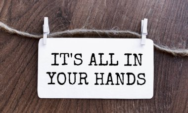 The text IT IS ALL IN YOUR HANDS is written on a piece of paper with clothespins attached to the winding. View from above. The badge with the inscription lies on a wooden table. Creative lettering.
