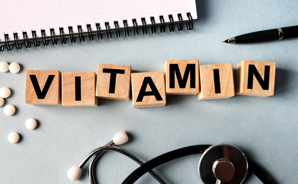 Wooden cubes with the inscription VITAMIN lie on the table. View from above. Nearby is a stethoscope, a notebook and scattered pills.