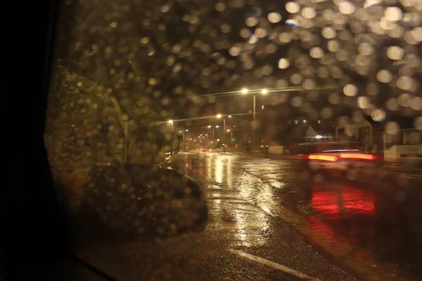 Shot taken through the car window of a night scene on wet cold night near the beach, street lights shinning and cars driving past, the rain drops on the car window reflecting the light. red lights of car driving past.