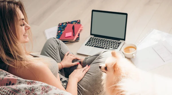 Genç bir kız köpeği Pomeranian ile yerde oturuyor ve bir dizüstü bilgisayar üzerinde çalışıyor. Köpek ona bakıyor.