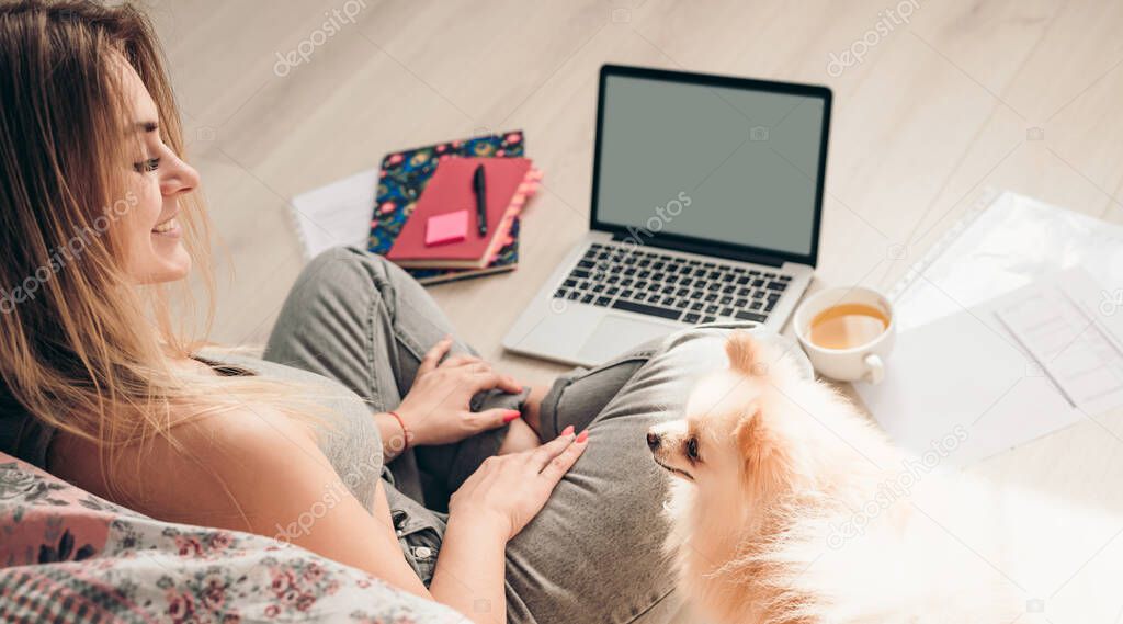 A young girl sits on the floor with her dog Pomeranian and works on a laptop. Dog is looking at her