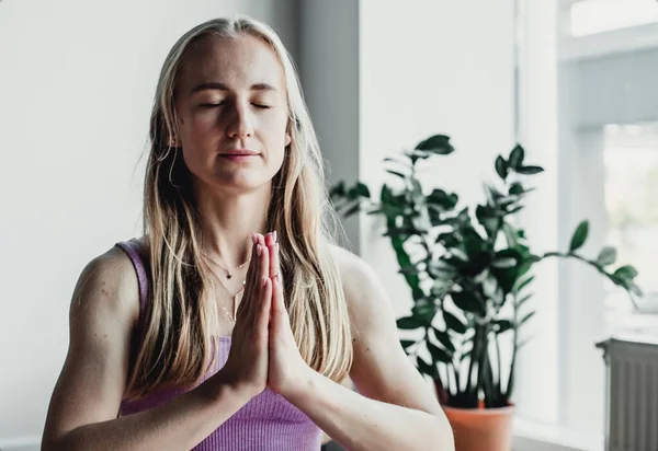 Genç çekici bir kadın yoga yapıyor Padmasana 'da oturuyor, egzersiz yapıyor, Lotus pozu veriyor, namaste yapıyor spor yapıyor, spor kıyafetlerini giyiyor, pencere kenarında.