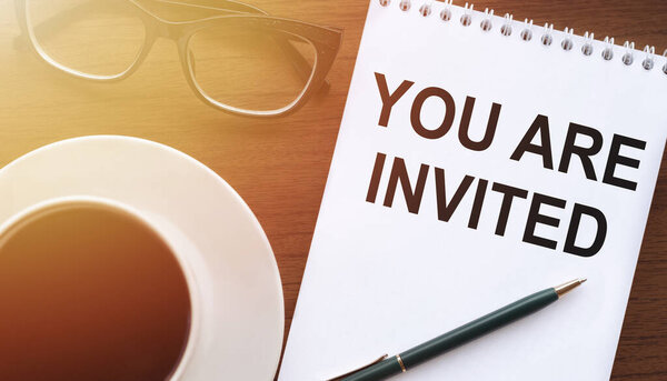 Текст "Вы приглашены" написан в блокноте, на столе с кофе, стаканами и ручкой.