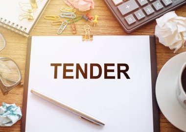 Ahşap bir masanın üzerinde TENDER yazılı bir ofis kağıdı var. Hesap makinesi, bardak, kalem, buruşuk kağıt ve bir fincan kahve ile iş yeri.
