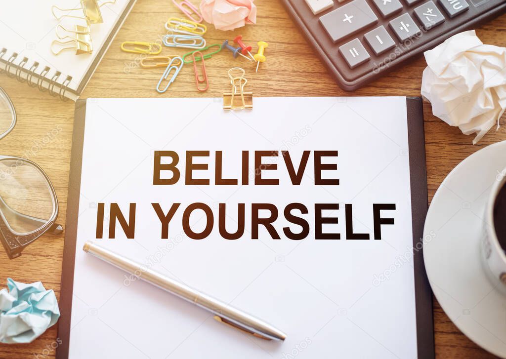 On a wooden table there is an office sheet of paper with the text BELIEVE IN YOURSELF. Business workspace with calculator, glasses, pen, crumpled paper and cup of coffee.
