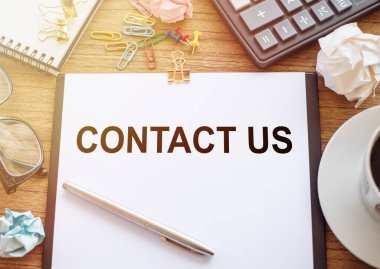 Tahta bir masanın üzerinde CONTACT US yazılı bir ofis kağıdı var. Hesap makinesi, bardak, kalem, buruşuk kağıt ve bir fincan kahve ile iş yeri.