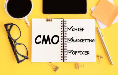 CMO - Pazarlama Şefi Kısaltma, not defterinde yazı ve sarı masa üzerinde ofis aksesuarları.