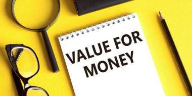 Notepad with the text VALUE FOR MONEY on a yellow background with glasses, a magnifying glass and a pencil. Business concept.