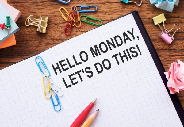 Notepad with the text HELLO MONDAY, LET\'S DO THIS on the table with colored paper clips and crumpled sheets.