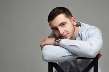 Mavi gömlekli dalgın esmer adam sandalyede oturuyor ve gri arka plandaki kameralara bakıyor.