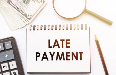 Geç ödeme metninin olduğu beyaz not defteri dolar ve ofis araçlarının arka planı.