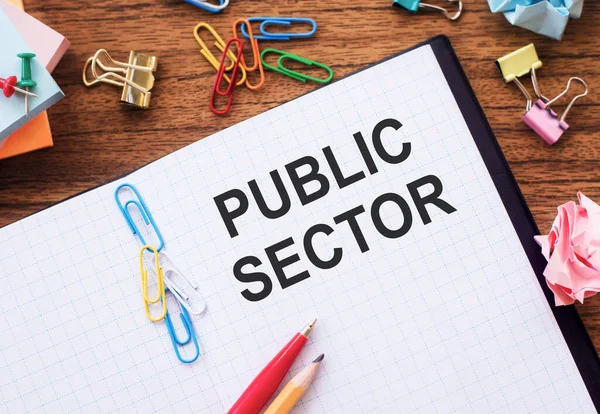 Notepad with the text PUBLIC SECTOR on the table with colored paper clips and crumpled sheets.