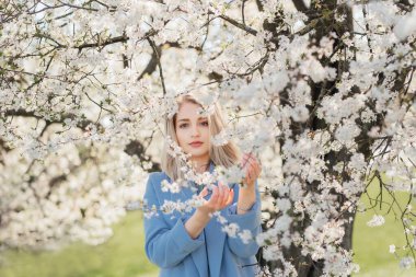 Güzel bir kız çiçekli bir ağacın altında duruyor ve elinde çiçeklerle bir dal tutuyor.