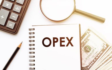 Dolar ve ofis araçlarının arka planı üzerinde OPEX (İşletim Harcamaları) metni bulunan beyaz not defteri.