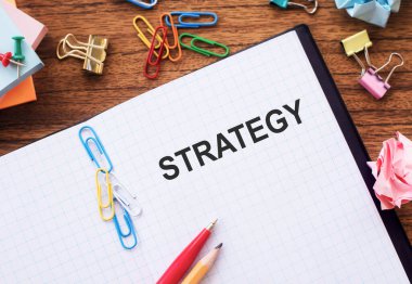 Masanın üzerinde renkli ataçlar ve buruşuk kağıtlarla STRATEGY metni olan not.