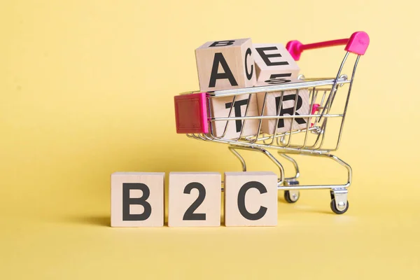 The word B2C - Business-to-consumer, on wooden cubes, on a yellow background with a shopping trolley.