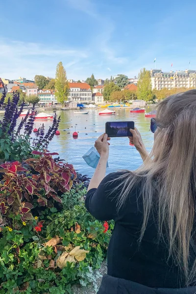 Blonde Frau beim Fotografieren mit ihrem Smartphone in der Bucht von Lausanne. Chirurgische Maske in ihrer Hand — Stockfoto