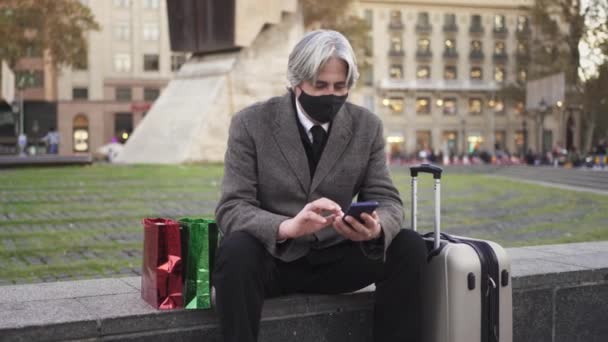 Людина з масками обличчя, подарунками і валізою повертається додому в день Різдва Коронавірусу. Старший подорожній розмовляє по телефону. — стокове відео