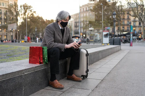 Man with face mask, presents and suitcase is going back home in Coronavirus Christmas season. Senior traveler using phone
