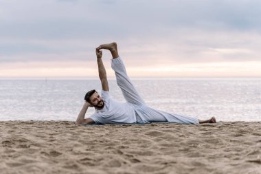 Beyaz elbiseli, sakallı modern sağlıklı adam plajda yoga egzersizleri yapıyor.