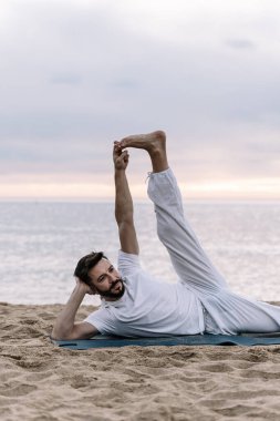 Beyaz elbiseli sakallı bir adam kumsalda yoga egzersizleri yapıyor.