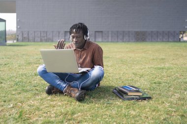 Genç zenci Afrikalı erkek öğrenci bilgisayar ve kulaklık kullanarak kampüsün dışında kitap ve defterlerin yanında çimlerin üzerinde oturuyor.