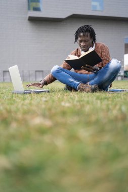 Afrikalı öğrenci kitap okuyor ve dizüstü bilgisayarla üniversite projesi için bilgi topluyor, kampüsün çimlerinde.