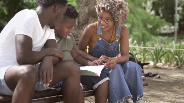 Fiatal Y-generációs fekete szülők segítik gyermeküket az iskolai házi feladat elkészítésében a parkban. — Stock videók