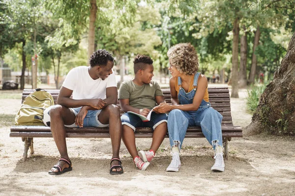 Jeg ser svart familie gjøre skolelekser på en benk i parken. Liten sønn som gjør skolearbeid med hjelp av foreldrene – stockfoto