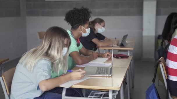 Gimnazjaliści z maską ochronną zapisujący notatki podczas lekcji w klasie liceum — Wideo stockowe