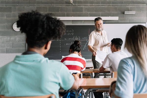 Mature friendly professor with beard giving corrected exams sheets to asian student in class. Rear view of multi-ethnic teenager students in college high school classroom. Teacher returning test