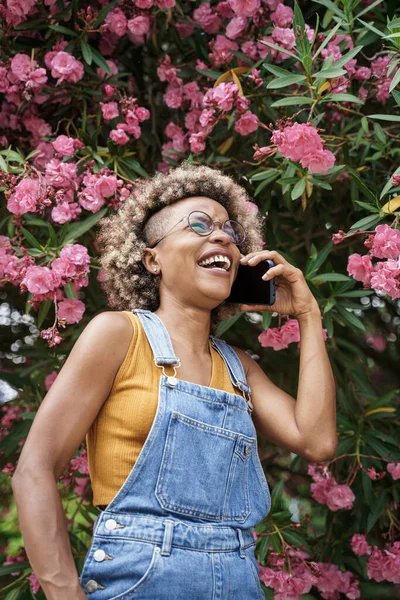 Afrikkalainen amerikkalainen tyylikäs nainen hymyilee puhelun ympärillä vaaleanpunaisia ruusuja puutarhassa — kuvapankkivalokuva