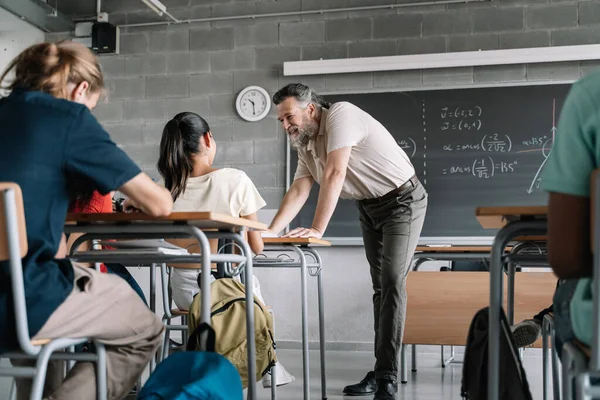 Hyggelig lærer med skjegg som hjelper elever i matte naturfag. Professoren snakker med jenta i klasserommet. – stockfoto