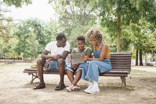 Afrikkalainen amerikkalainen perhe hauskaa tabletin katsella online-videoita ja sosiaalisen median. Nuori viileä tuhatvuotinen vanhemmat ja söpö hymyilevä lapsi istuu penkillä ulkona julkinen puisto — kuvapankkivalokuva