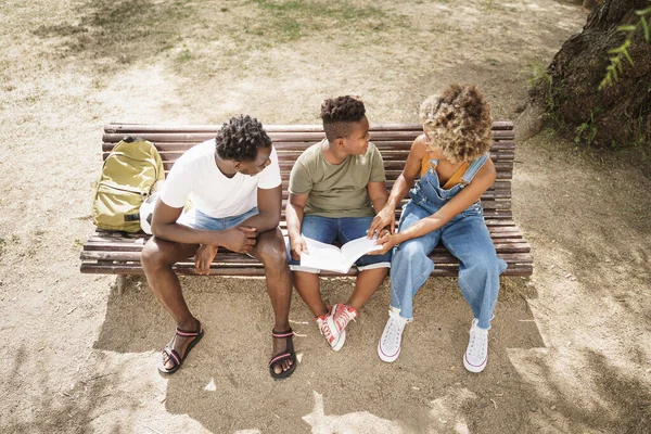 Toppkarakter på afroamerikansk familie som gjør lekser på en benk i parken. Liten sønn som gjør skolearbeid med hjelp av foreldrene – stockfoto