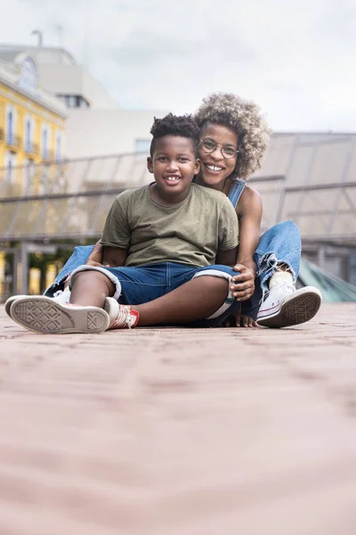 Smilende svart gutt lener seg mot mor som sitter tett sammen på byens gate. Portrett av lykkelig afroamerikaner som elsker mamma og sønn utenfor – stockfoto