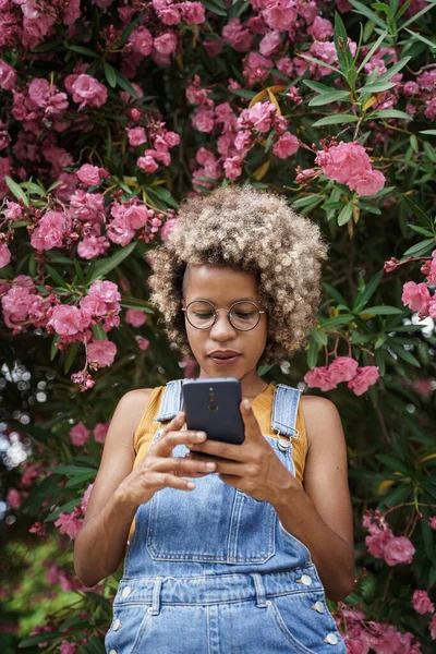 Cool nuori moderni musta nainen vaaleat hiukset älypuhelimella sosiaalisen median ympäröivät vaaleanpunaiset ruusut puutarhassa — kuvapankkivalokuva