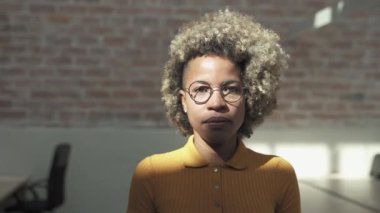 Şık, neşeli ve kendine güvenen Afro-Amerikan genç kadın portresi. Sarı saç kesimi, gözlükler ve tuğla duvarlı modern bir ofiste günlük giysi.