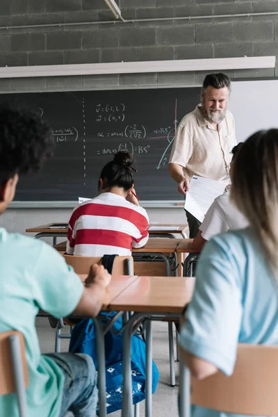 Lærer med skjegg som gir matematikk til en elev i timen. Synsvinkel på multietniske tenåringsstudenter i klasserom for videregående skoler – stockfoto