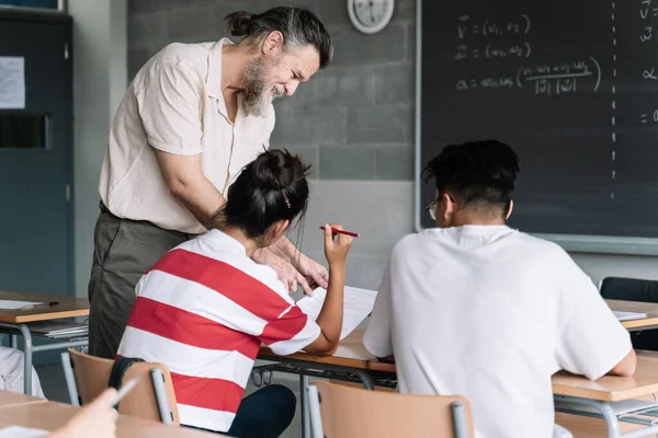 Vennlig professor som forklarer matematikkeksamen for tenåringsstudenter i klasserommet. Seniorlærer med skjegg hjelper student – stockfoto