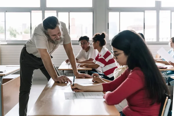Vennlig professor som forklarer øvelsen til elevene i klasserommet. Seniorlærer med skjegg hjelper tenåringsstudent – stockfoto