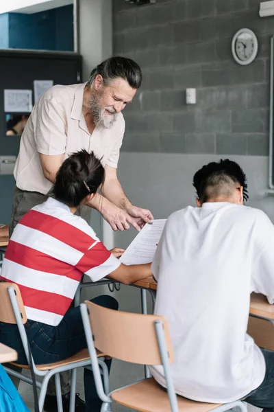 Seniorlærer med skjegg hjelper student. Vennlig professor som forklarer eksamensøvelse for tenåringsstudenter i klasserommet – stockfoto