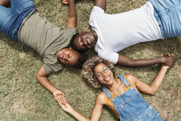 En munter svart familie som holder hender mens de ligger på gress sammen i en park. Lykkelige unge afroamerikanske svarte foreldre som elsker sønnen sin – stockfoto