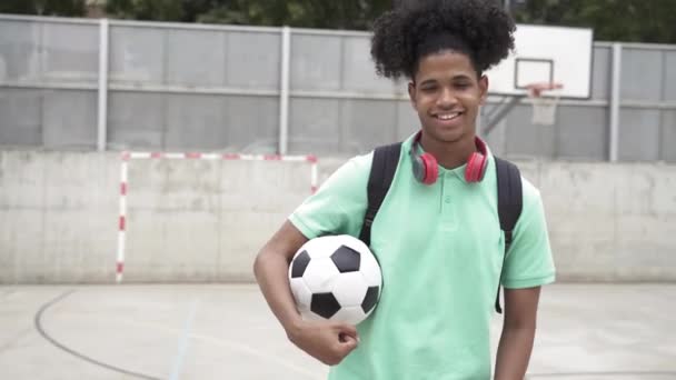Portrait d'un jeune étudiant noir afro-américain souriant avec chemise verte, écouteurs rouges et ballon de football dans la cour de basket du lycée — Video