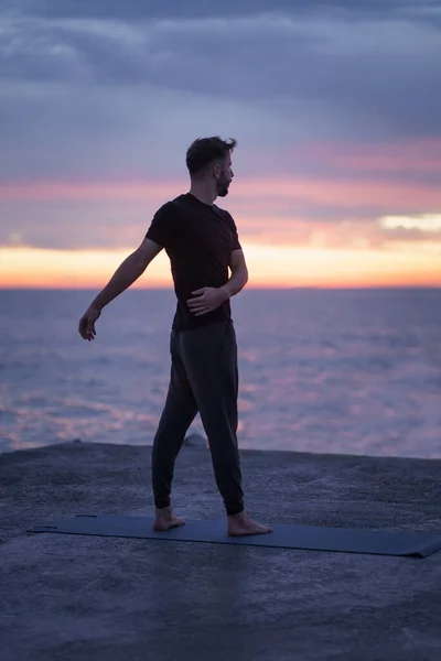 Mies venyttely ja tekee terve aamu wellness rutiini meren rannalla edessä auringonnousun dramaattinen taivas — kuvapankkivalokuva