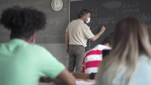 Nauczyciel i nastolatek Uczniowie z maską twarzy uczenia się w klasie liceum. Profesor piszący formuły matematyczne na tablicy — Wideo stockowe